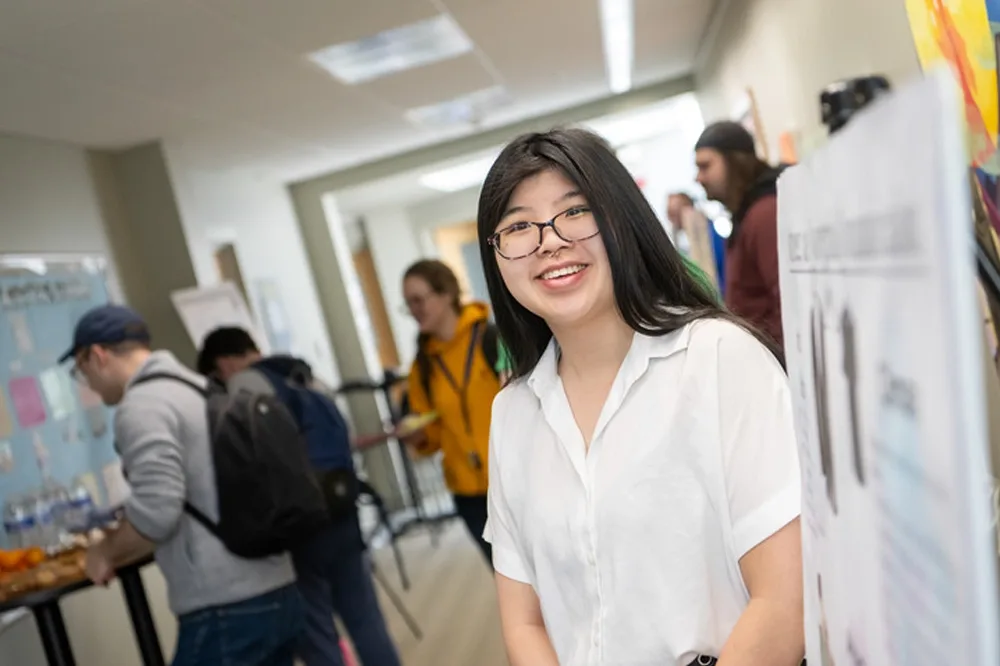 Student wearing glasses standing next to a capstone presentation poster in a hallway.