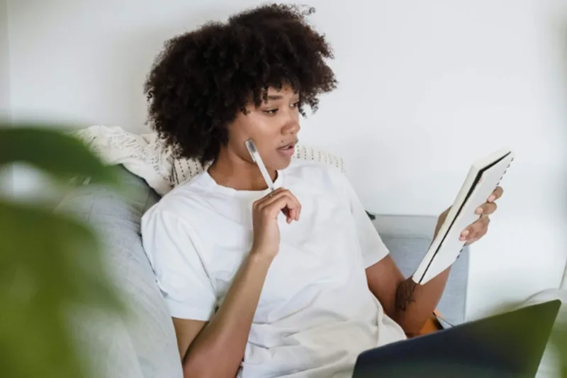 A woman reading a notebook while holding a pen up to her cheek.