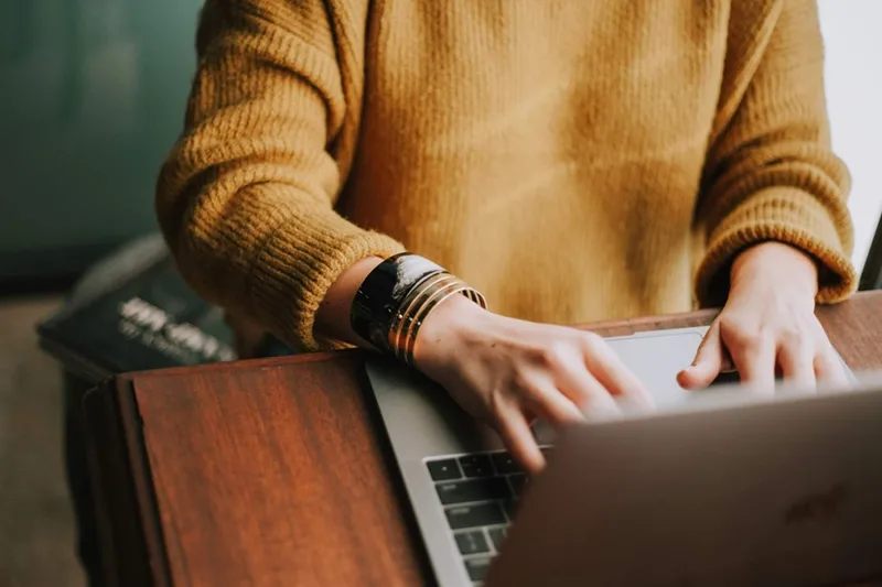 An individual wearing a gold sweater typing on a silver laptop.