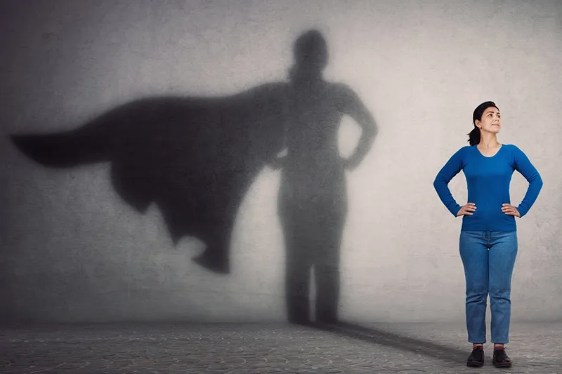 A woman in a blue shirt, with her shadow displaying a superhero cape on the wall behind.