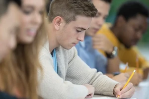 People sit at a table with laptop and pencils. Four people are out of the focus, with the fifth person, in the center, in focus and holding a pencil.