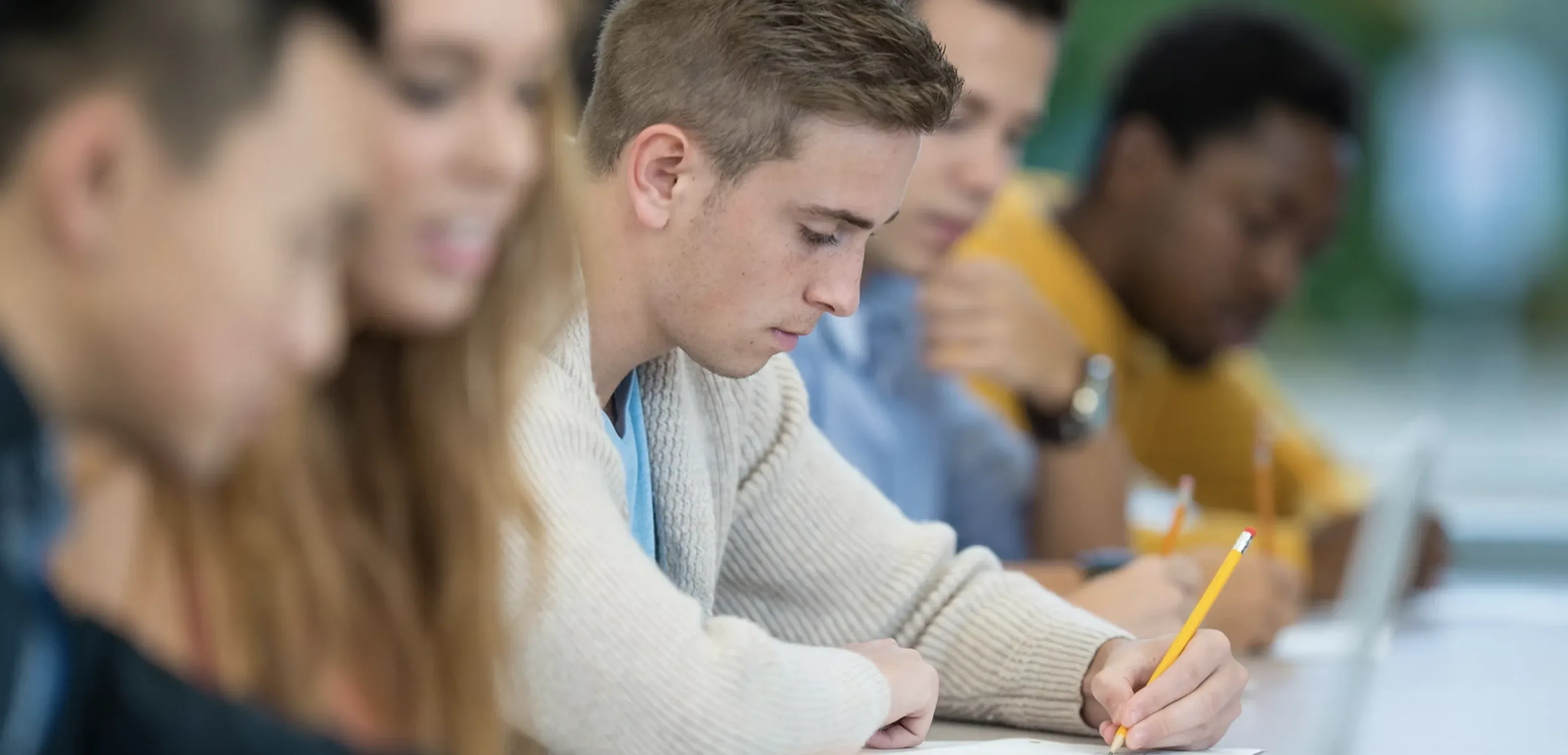People sit at a table with laptop and pencils. Four people are out of the focus, with the fifth person, in the center, in focus and holding a pencil.