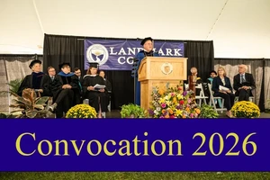 People sit on a stage on the Landmark College quad at a previous convocation. At a podium is College President Jim Dlugos in a cap and gown. Behind Dlugos is a dark blue banner that says "Landmark College" with the College logo. A blue graphic goes across the bottom of the screen that reads "Convocation 2026" in yellow letters.