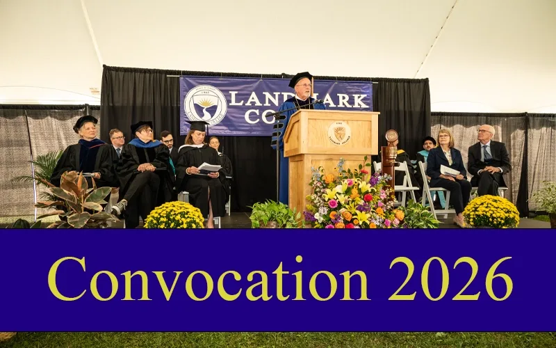 People sit on a stage on the Landmark College quad at a previous convocation. At a podium is College President Jim Dlugos in a cap and gown. Behind Dlugos is a dark blue banner that says "Landmark College" with the College logo. A blue graphic goes across the bottom of the screen that reads "Convocation 2026" in yellow letters.