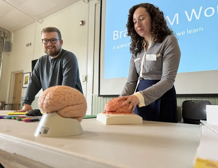 Two people present at a table with a projector screen behind them. On the table is a sculpture of a brain.