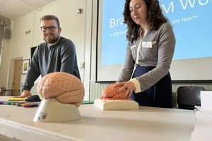 Two people present at a table with a projector screen behind them. On the table is a sculpture of a brain.