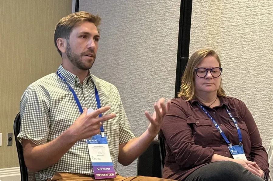A man and woman wearing lanyards and badges sit in chairs. The man is talking and gesturing.
