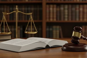Symbolic scales of justice, a book, and a gavel are see on a wooden table. In the background is a bookshelf.
