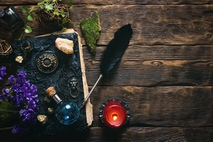 A spellbook, ring, potion, feather, plant, flowers, ball of tweed and a candle are on a wooden surface.