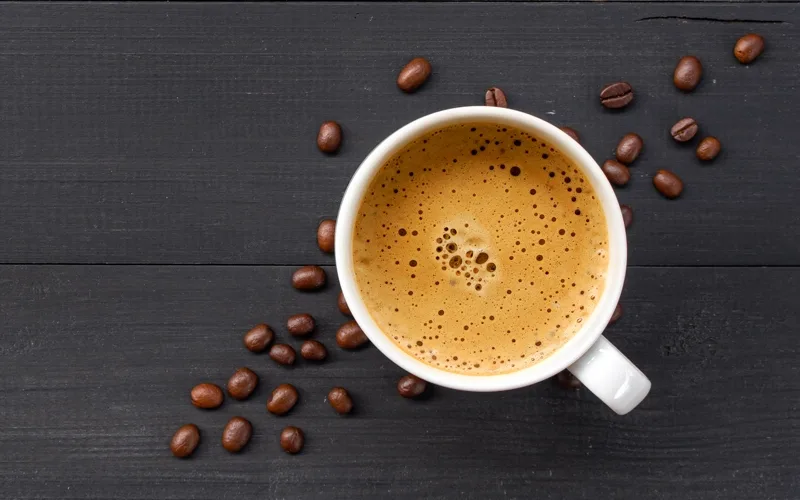 An aerial view of a cup of coffee with coffee beans scattered on a gray wooden surface.