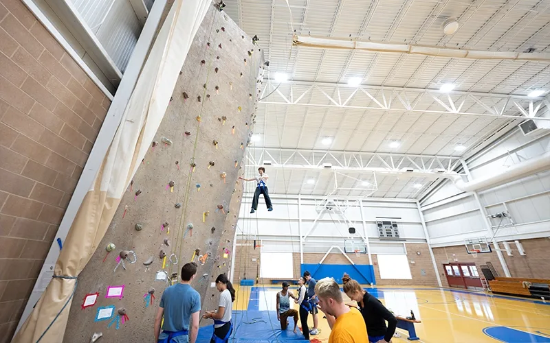 A group of people hangs out and climbs the climbing wall at Landmark College.