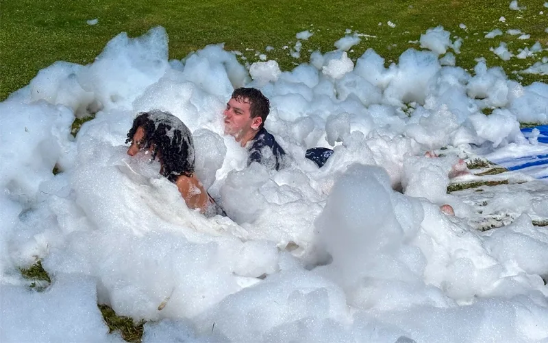 Two people have just slid into a pile of bubbles at the end of a slip-and-slide. Their faces are covered in bubbles.