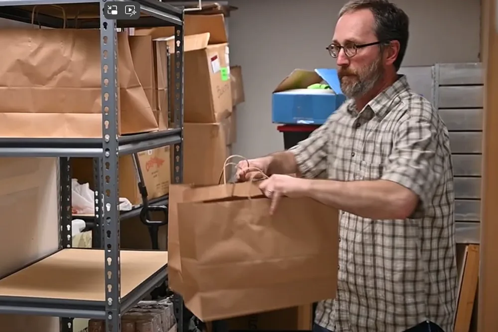 Man with glasses in storage room holding two brown shopping bags.