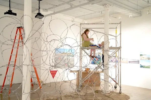 Yishay Garbasz sitting on scaffolding in an art gallery, assembling a barbed wire sculpture.