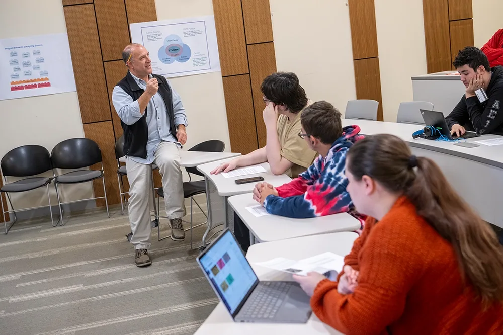 A professor with a yellow pencil behind his ear, talking to a class full of students.