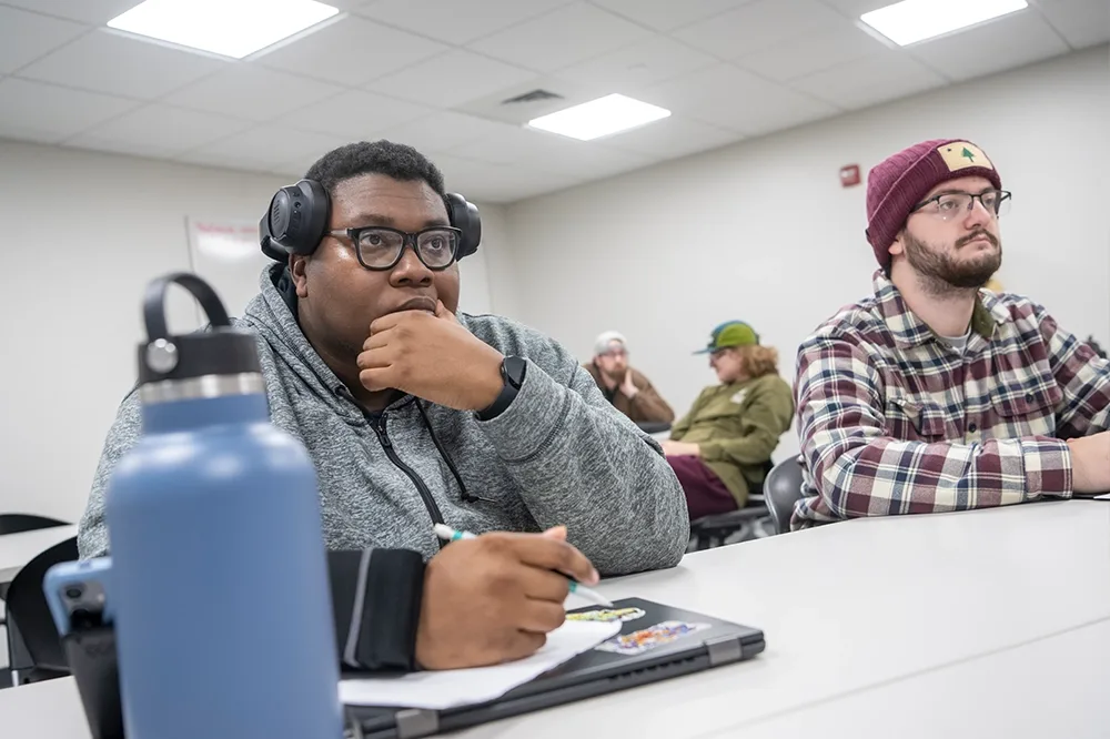 A student taking notes with a blue water bottle in front of him beside a student in a plaid shirt and hat sitting to the right.