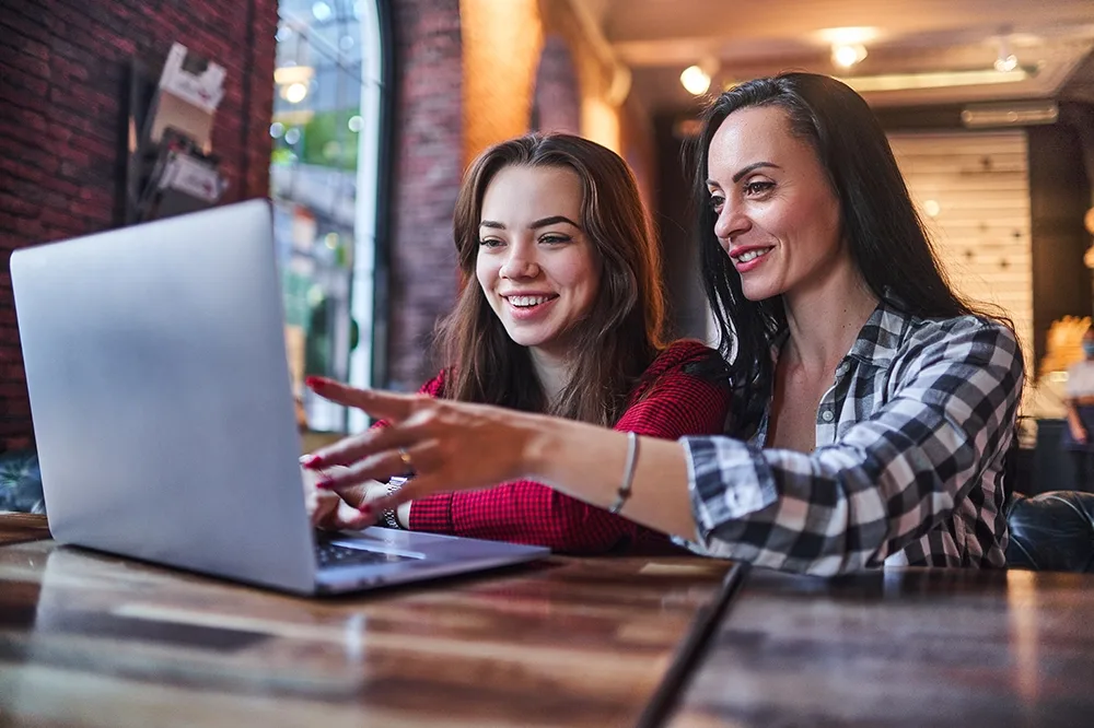 A mother with her teen daughter sitting in front of a laptop smiling and pointing to the screen.