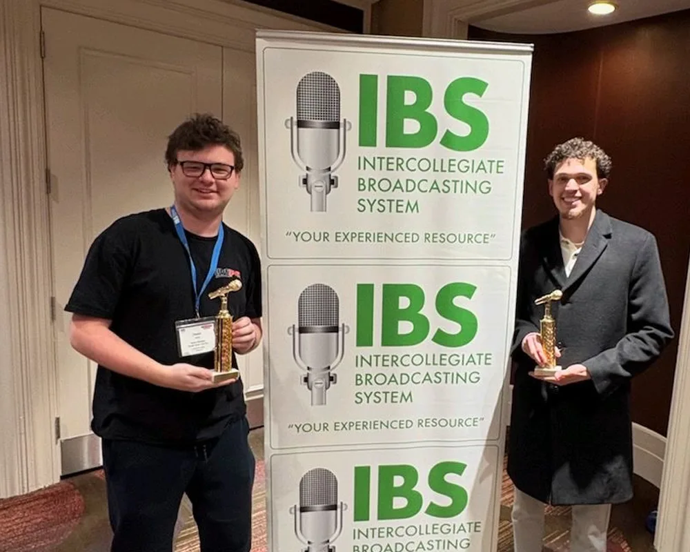 Two students holding their award trophies while standing next to an Intercollegiate Broadcasting System pull-up banner.