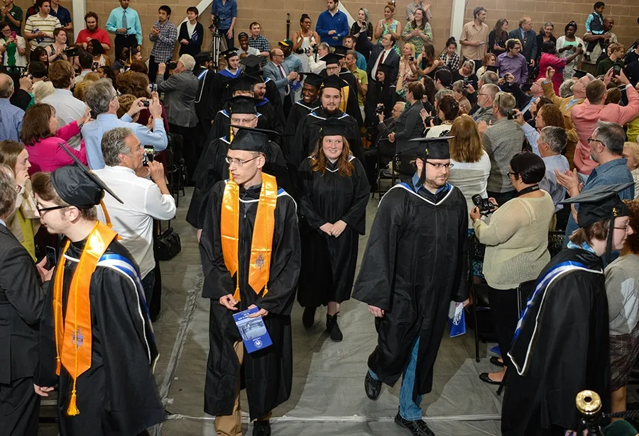 People in caps and gowns process down the middle, with onlookers on either side taking photos.