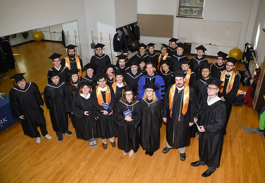 People in caps and gowns stand on a finished wood floor next to a mirror.