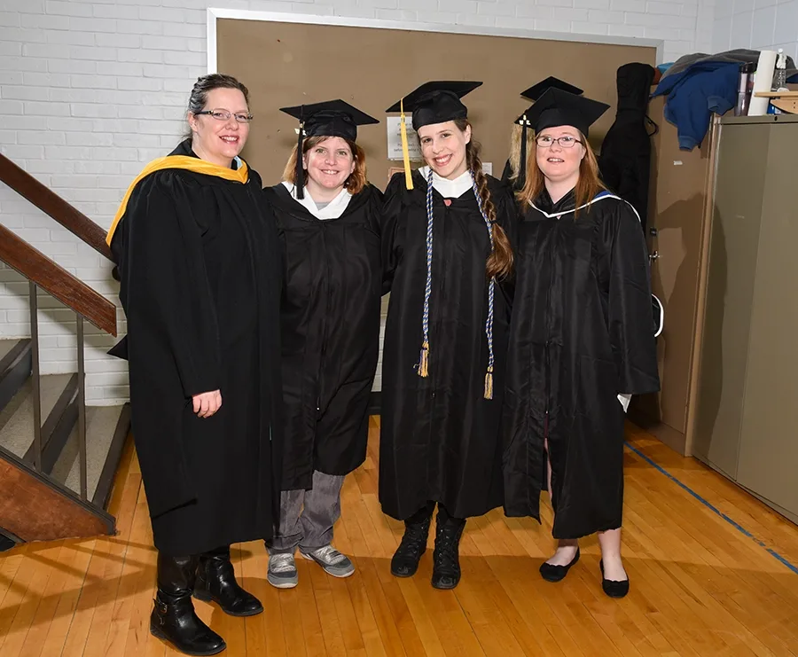 Four people pose with their arms around one another for a photo. The first individual is wearing a gown with a yellow sash. The next three are wearing black caps and gowns.