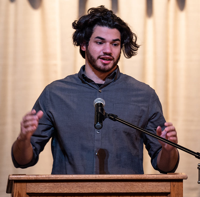 Male with long wavy black hair and beard talking at podium