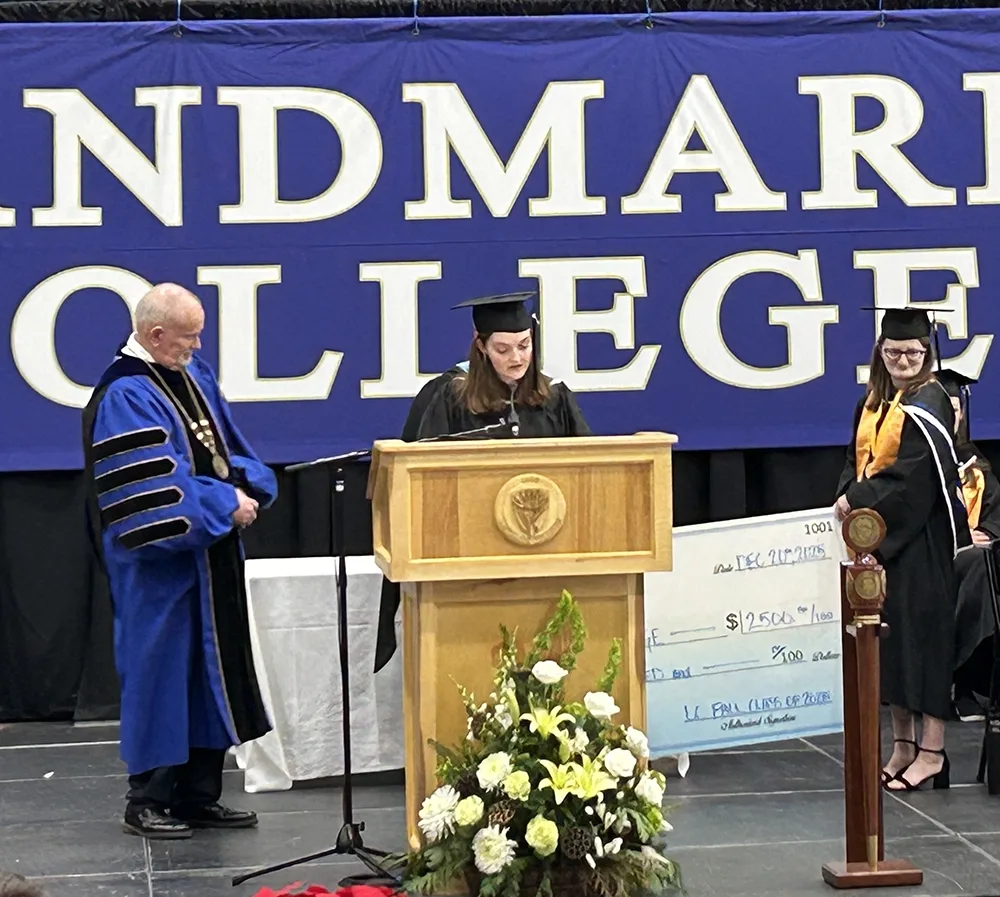 Woman speaking at podium with man to her right listening and woman to her left holding an oversized check. All are wearing graduation regalia