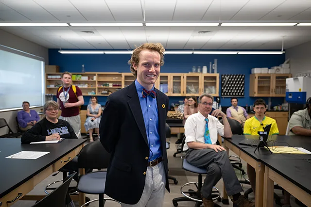 Dylan Sullivan posing with a bowtie and blue shirt in a classroom with the professor and students behind him.