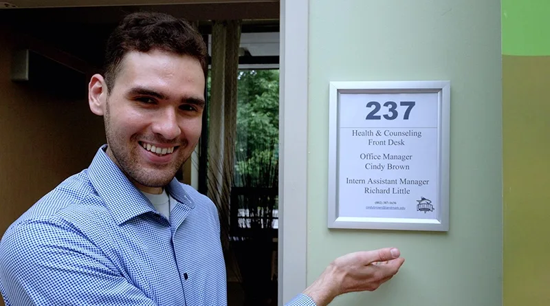 Richard Little, a male student with short brown hair and wearing a blue shirt, smiles and gestures at a sign that has his name and title of intern assistant manager.