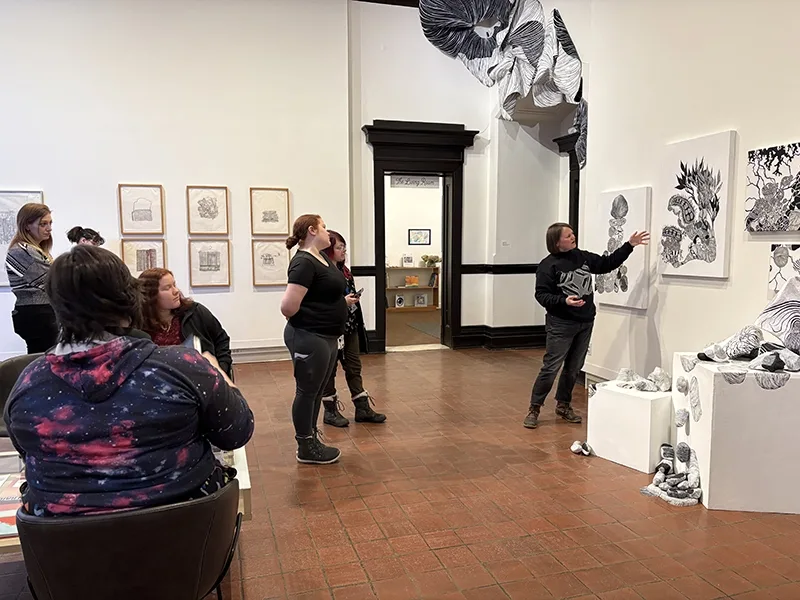 A woman gestures at black and white art hanging on a wall in a large gallery space with several students looking on