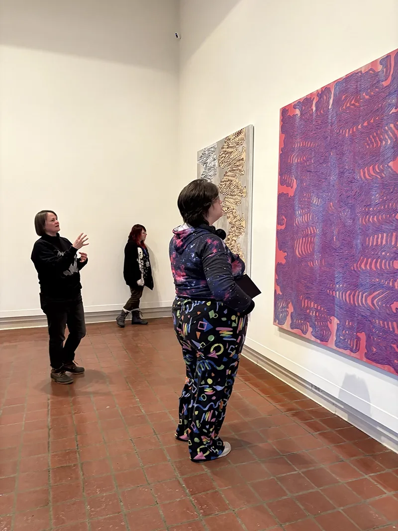 Student looking at large colorful artwork on a gallery wall with a woman standing behind her and gesturing in explanation.