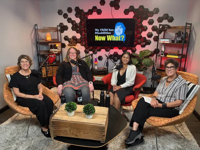 Four Staff members sitting in a room with a monitor on the wall displaying Now What?