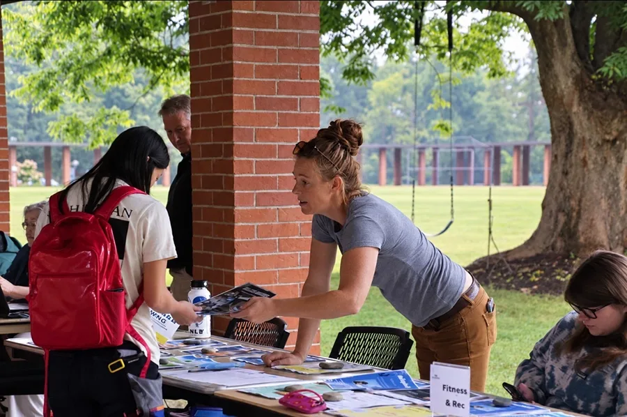 A person at a table promoting "Fitness & Rec" activities hands a flyer to someone looking on.