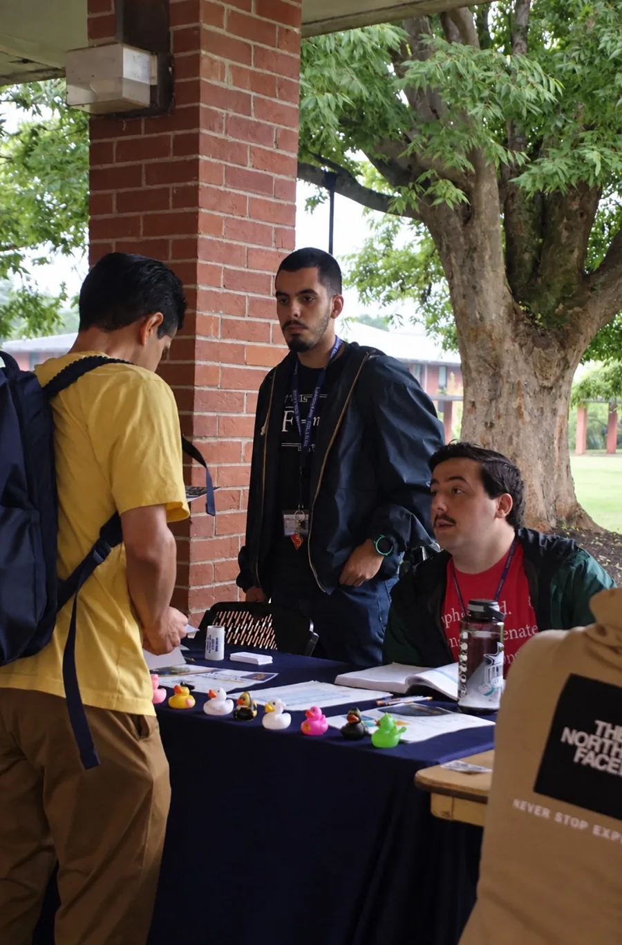 Two people stand behind a table promoting a campus activity while one person looks on. On the table is a row of colorful rubber duckies.