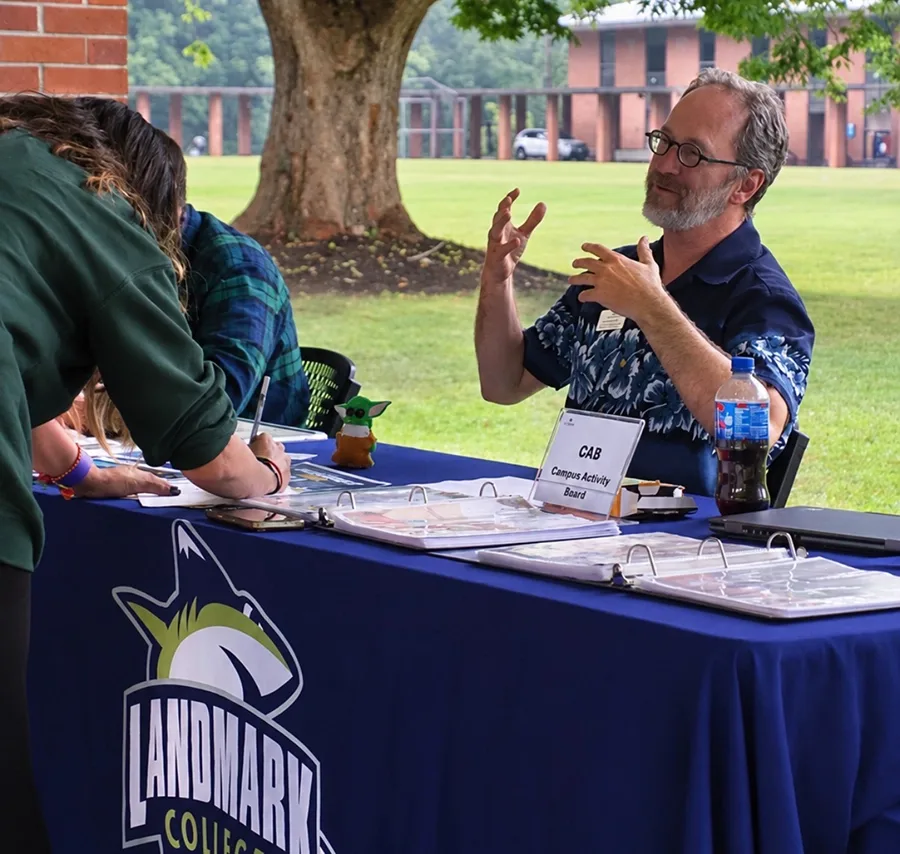 A person talks with his hands at a table labeled "Campus Activity Board" while another person signs a sign-up sheet. There is a small green Yoda-like figurine on the table. A blue tablecloth displays the Landmark College logo of a shark and the words "Landmark College."