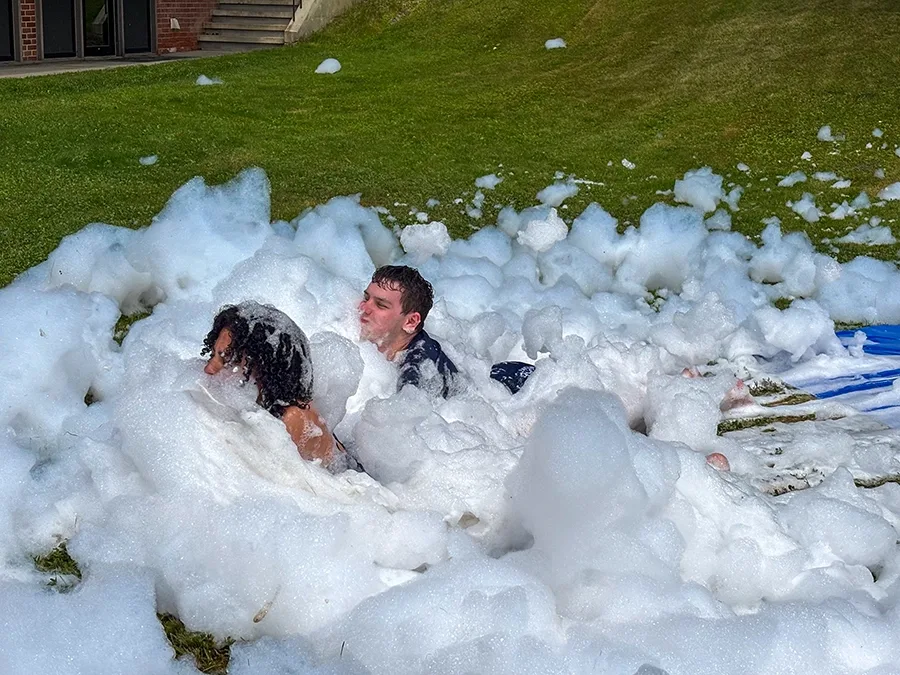 Two people have just slid into a pile of bubbles at the end of a slip-and-slide. Their faces are covered in bubbles.