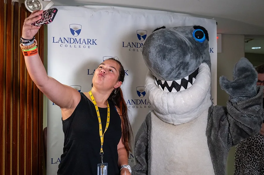 A person holds up their phone while making a silly face to take a selfie with Finn, a shark mascot.