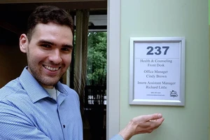 Richard Little, a male student with short brown hair and wearing a blue shirt, smiles and gestures at a sign that has his name and title of intern assistant manager.