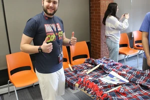 Male student in shorts and t-shirt giving thumbs up signal with hands as he stands next to the no sew blanket he is assembling.