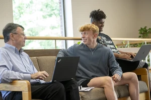 A professor and two male students sitting together on a couch and laughing about something.