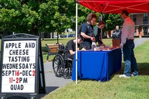 Some people at an apple and cheese tasting booth. In the foreground is a standing sign that reads: "Apple and Cheese Tasting Wednesdays Sept 16 - Dec 2 11 - 2 p.m. Quad"