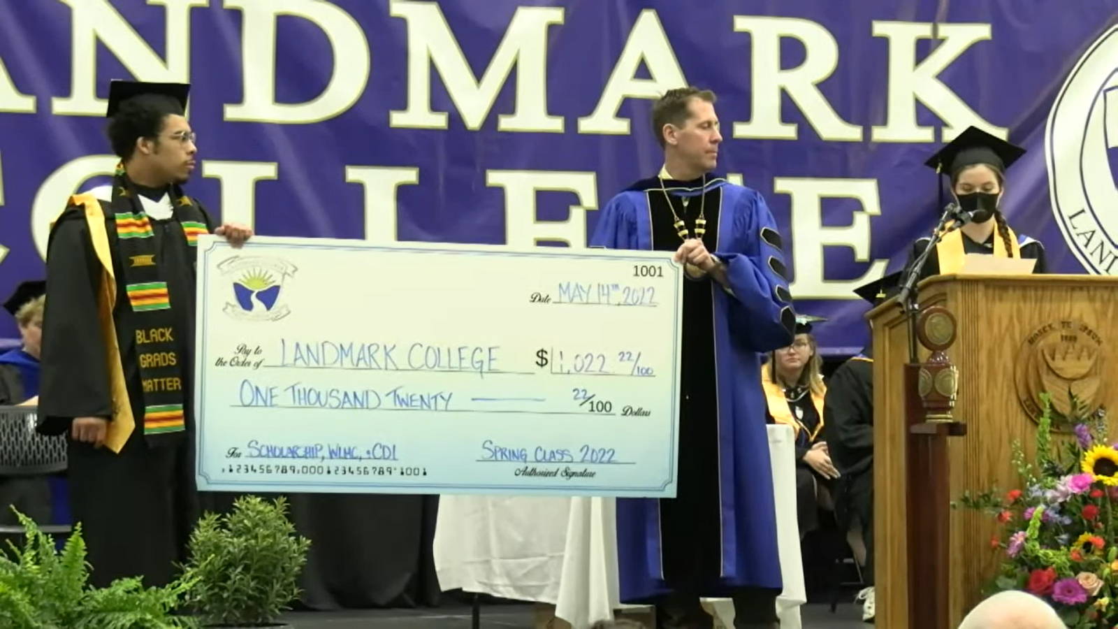 Student Carter Griffith and President Peter Eden, both in graduation regalia, standing on the commencement stage holding an oversized check in the amount of 1,022.22. They are both looking to their left  at student Meredith Robertson, who is making remarks at the podium while wearing a protective mask.