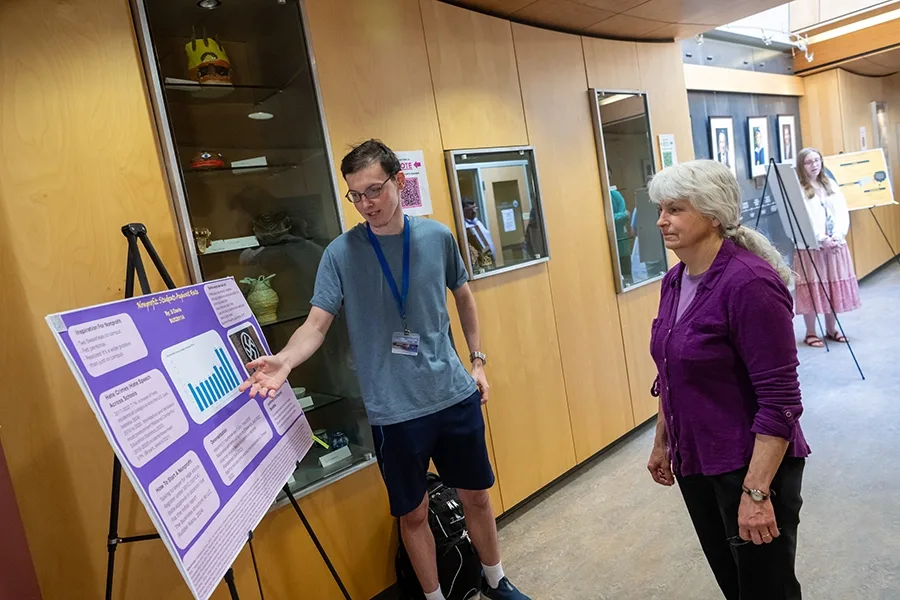 Student JJ Davis shows a presentation on a poster and easel to faculty member Lee Crocker in a hallway with other student presenters.