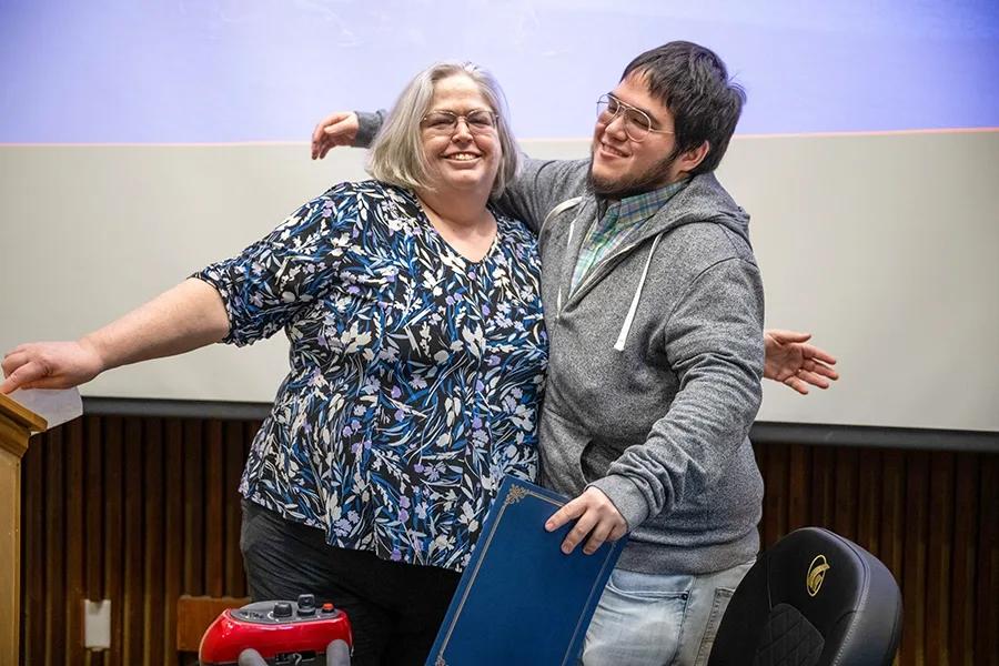 Faculty member Jeanette Landin and student Gabe Kwok hug each other at an award ceremony. Kwok is holding an award.