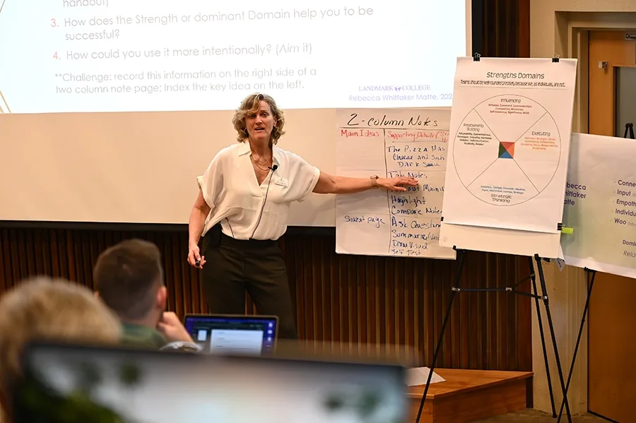 Faculty member Rebecca Matte stands at the front of a lecture hall gesturing to slides and diagrams as people look on and listen.