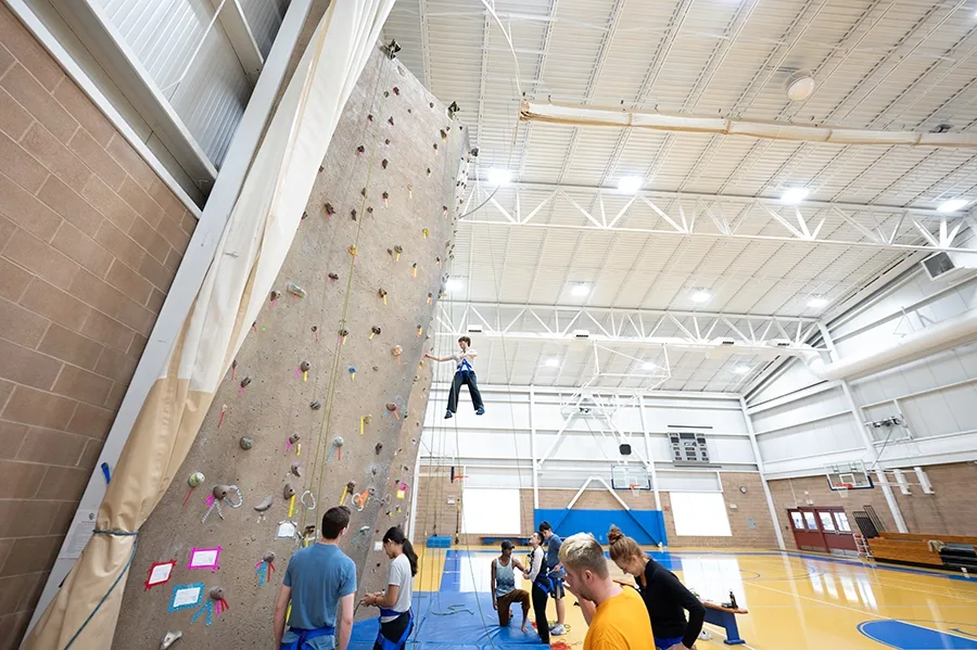 A person in a harness drops down from a climbing wall in the Landmark College Click Center while others wait at the bottom.