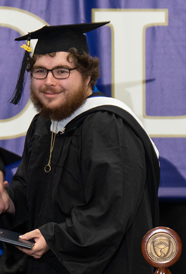 Man with bushy beard and dark rimmed glasses wearing black graduation cap and gown smiles while shaking hands with someone off camera.