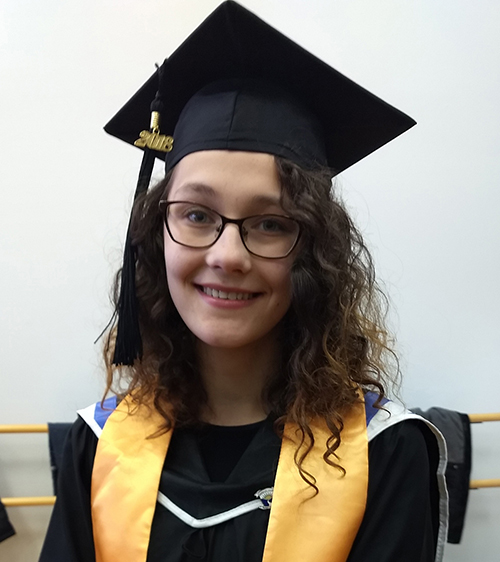 Katie LaBombard, a woman with long, curly brown hair and glasses. She is dressed in a black graduation cap and gown with a gold honors sash.
