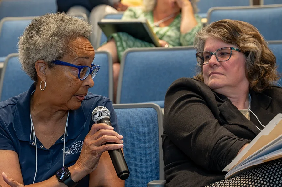 Two people sitting in an auditorium are looking at each other. One of them is speaking into a microphone.