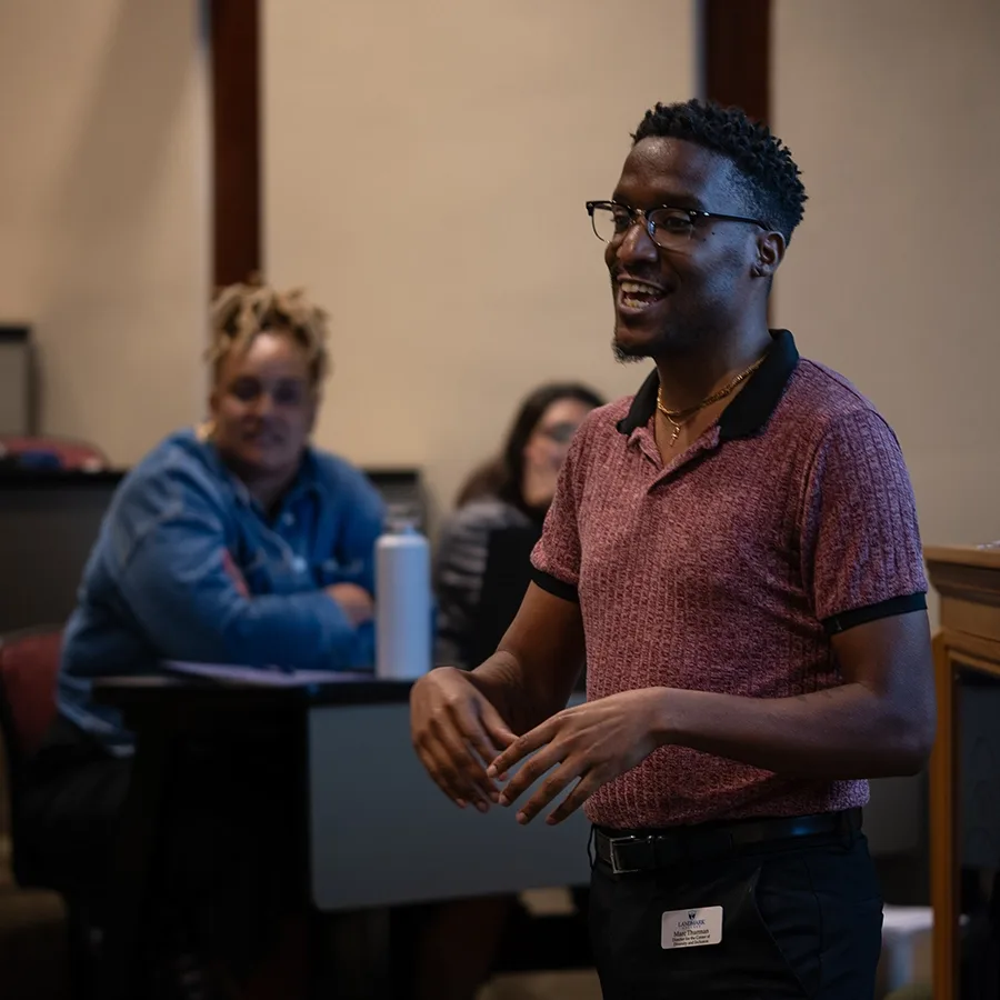 A person stands in a lecture hall and speaks. Two people are shown seated in the foreground.